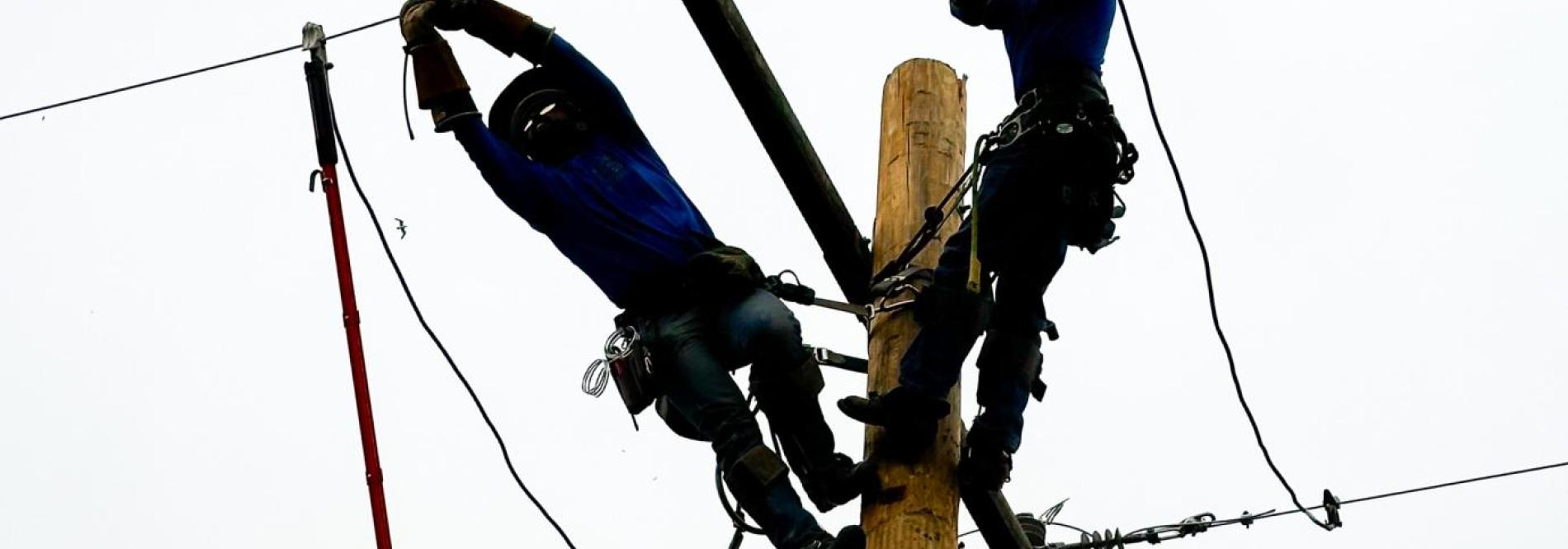 Two electrical lineworkers perform maintenance work on top of a power pole
